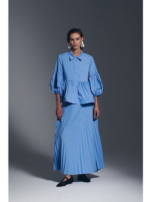 A woman models the MILVA MI Cotton Pleated Skirt in Azzurro Blue by MILVA, paired with a matching flowy top and voluminous sleeves, styled with bold white earrings against a plain gray background.