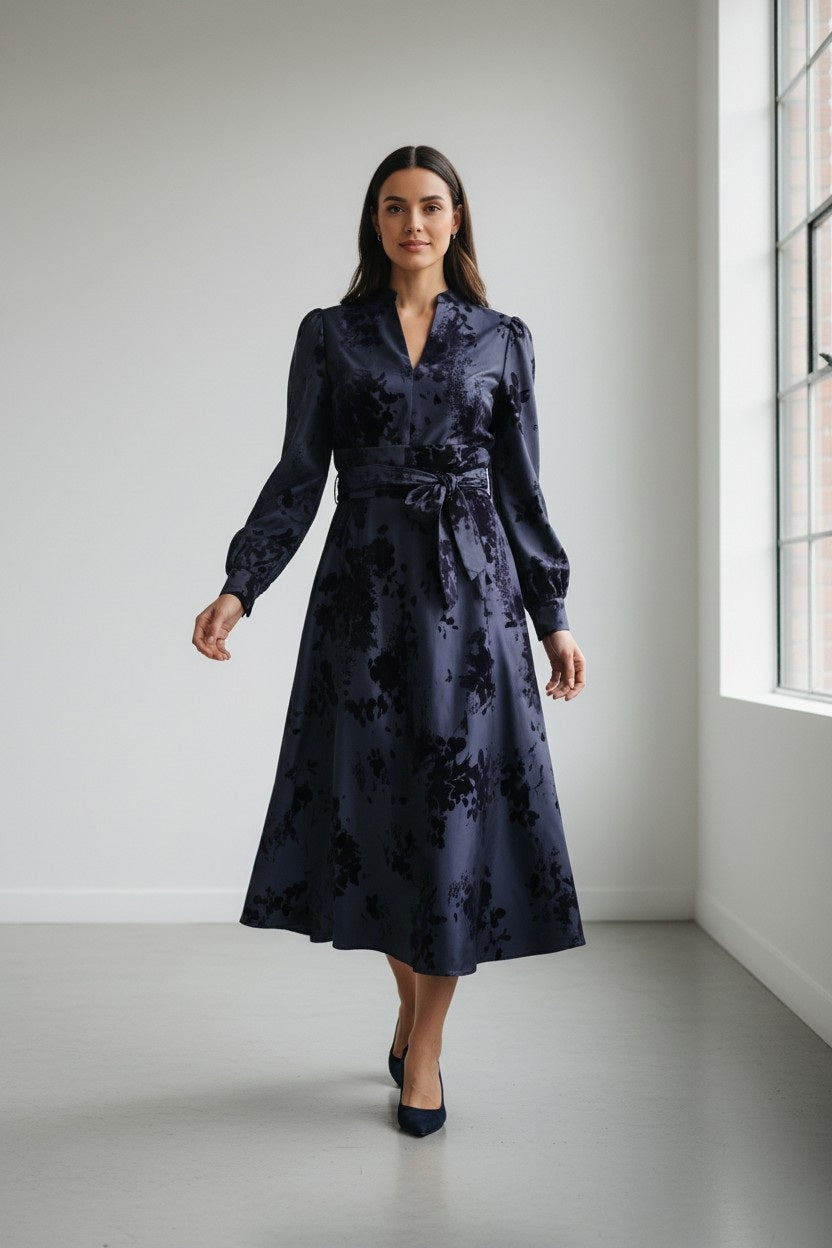 A woman stands indoors, facing forward near a large window in a bright, minimalist room. She is wearing the ROSSO35 Long Sleeve Taffeta Dress with Velvet in Navy Blue by ROSSO 35 SILKY—an elegant, belted midi dress featuring floral patterns and a luxurious printed flocked taffeta fabric, perfect for special occasions.