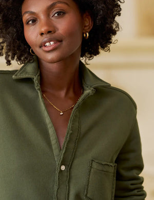 In a sunny, rustic kitchen, a woman smiles while wearing the FRANK & EILEEN EILEEN Relaxed Button Up in Triple Fleece Caper, paired with black wide-leg pants and sandals. She touches her hair as sunlight fills the cozy space.