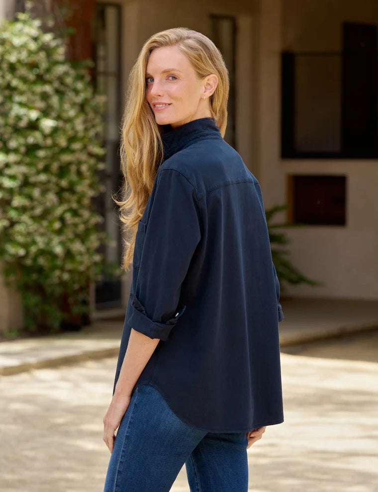 A woman stands in a minimal room, smiling with one hand in her pocket, wearing the FRANK & EILEEN - Waverley Classic Button Up Tee in Vintage Jersey Classic Navy from the California Heritage Collection, paired with blue jeans and brown ankle boots. A large window to the left fills the space with natural light.