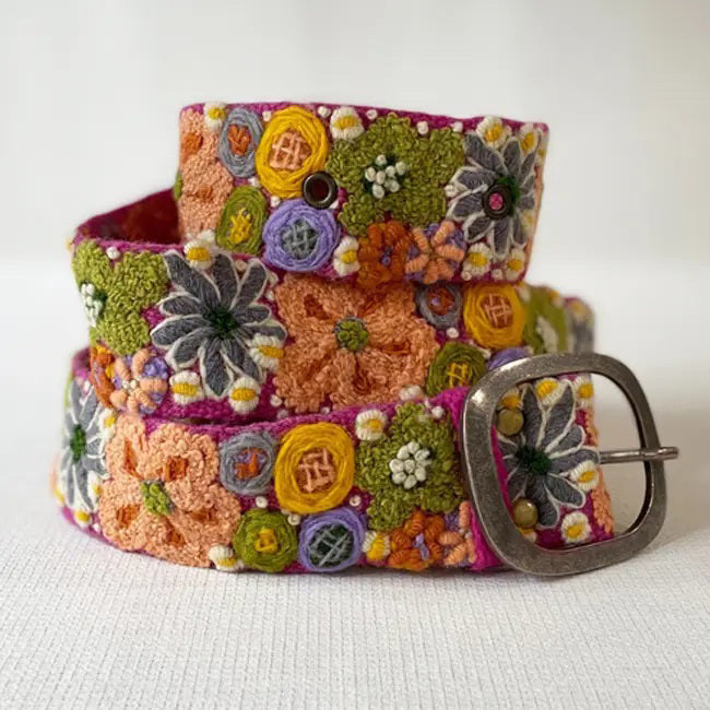 The JENNY KRAUSS WILDFLOWERS EMBROIDERED WOOL BELT features hand-embroidered floral patterns in orange, yellow, green, purple, and white on a peach background, crafted in Peru with a silver buckle and shown coiled on a white surface.