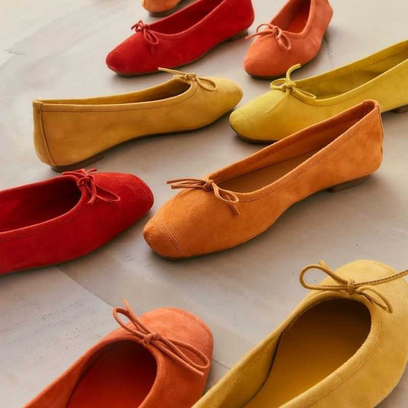 A person in white jeans, a beige coat, and carrying a round straw bag stands on light stone pavement, wearing REQINS HARMONY suede ballerina flats in rouge by REQINS.