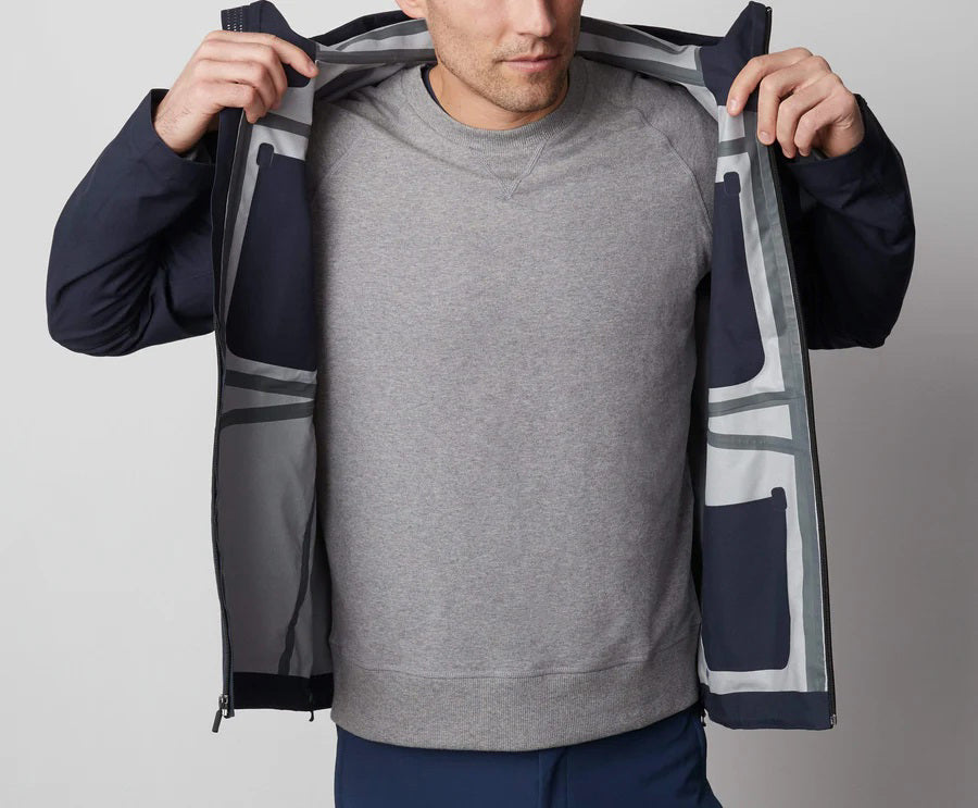 A man stands in front of a white wall wearing the MACK WELDON STORMCHASER JACKET with adjustable hood, paired with a gray shirt, blue pants, and black sneakers. He looks to the side while holding the jacket's zipper.