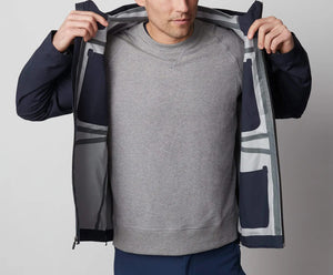 A man stands in front of a white wall wearing the MACK WELDON STORMCHASER JACKET with adjustable hood, paired with a gray shirt, blue pants, and black sneakers. He looks to the side while holding the jacket's zipper.