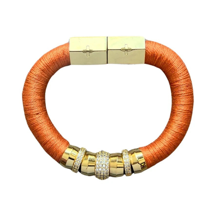 The HOLST + LEE - LUXE CLASSIC BRACELET by HOLST &amp; LEE features an elegant orange cord with gold-plated accents, including a rectangular gold clasp adorned with several gold and diamond-encrusted beads near the clasp.
