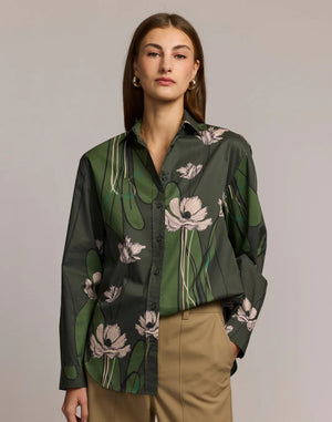 A woman with long brown hair wears the HINSON WU Halsey Long Sleeve Shirt in Poppy Print and tan pants, standing with her hands in her pockets against a neutral background.