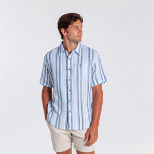A man wearing the CRIQUET - THE BEACHCOMBER SHIRT IN BLUE by CRIQUET, paired with light beige shorts, stands against a plain white background, looking to his left with one hand in his pocket.