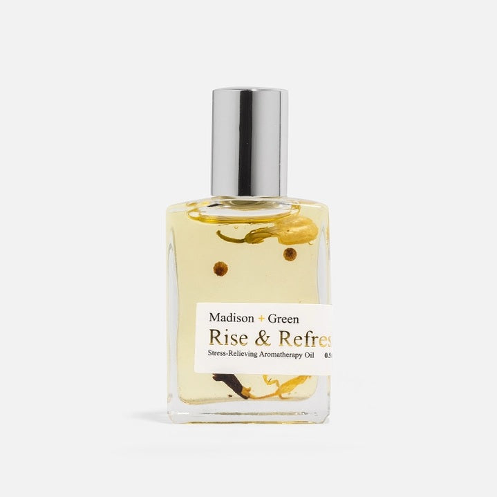 A small rectangular glass bottle labeled “MADISON AND GREEN - RISE & REFRESH MORNING AROMATHERAPY BODY OIL MINI” from MADISON GREEN, featuring a silver cap and visible botanical pieces suspended in the lightweight, plant-based oil.