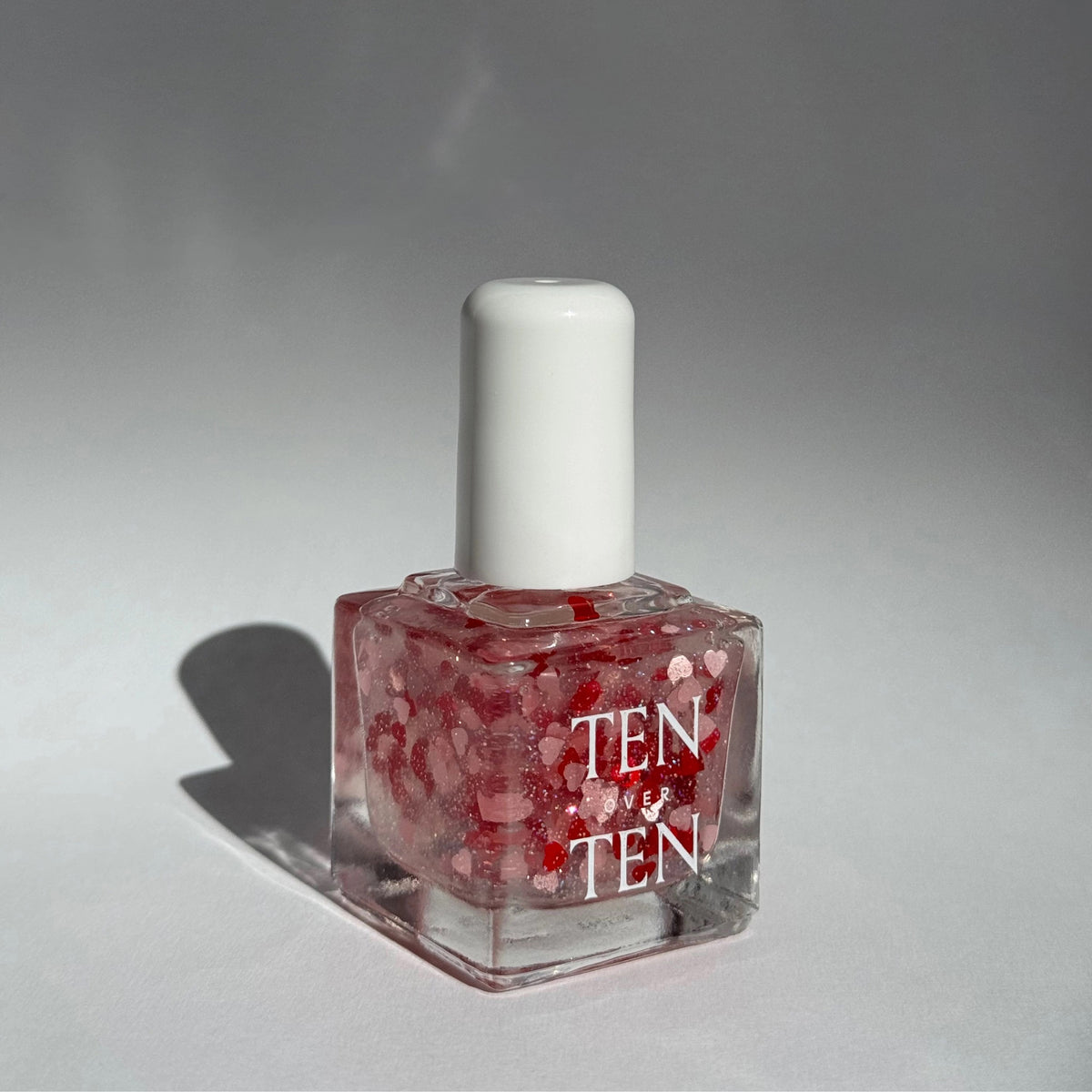 A glass bottle of TENOVERTEN - NAIL POLISH from the brand TEN OVER TEN, featuring a white cap and a dusty pink nail color. The front label displays "TEN OVER TEN" in white uppercase letters. This vegan, eight-free formula ensures both beauty and peace of mind. The background is plain and light-colored.