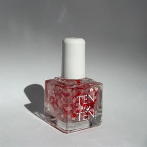 A glass bottle of TENOVERTEN - NAIL POLISH from the brand TEN OVER TEN, featuring a white cap and a dusty pink nail color. The front label displays "TEN OVER TEN" in white uppercase letters. This vegan, eight-free formula ensures both beauty and peace of mind. The background is plain and light-colored.