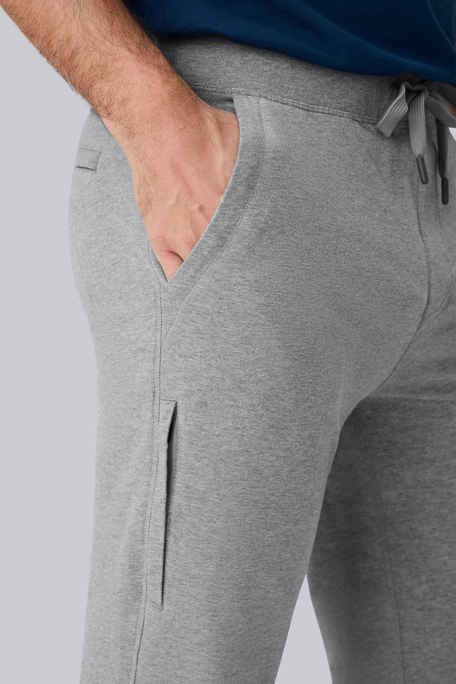A person wearing MACK WELDON ACE Sweatpants in Ice Storm Heather, a dark blue shirt, and gray sneakers with white soles stands on a light gray background, shown from the side with one arm relaxed.