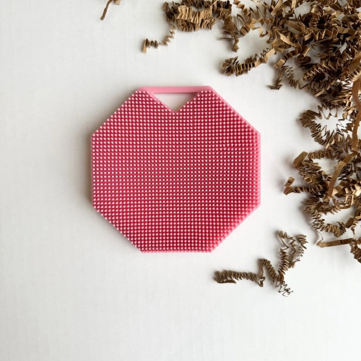 The SMILE BOUTIQUES SILICONE BODY AND EXFOLIATION BRUSH IN ROSE is an octagonal, pink silicone mat featuring small raised dots across its surface and a triangular cutout near the top edge, shown from above on a white background. This versatile brush serves as a reusable shower scrubber or body exfoliator, gently smoothing skin for a refreshed feel.