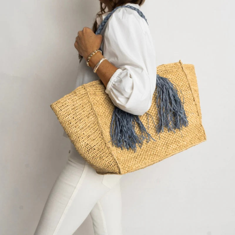 A woman in white shirt and pants holds the BALDIZ Camilla Raffia Tote Bag in Hay and French Blue, featuring chunky braided handles and long blue tassels—a handcrafted accent against a light background.