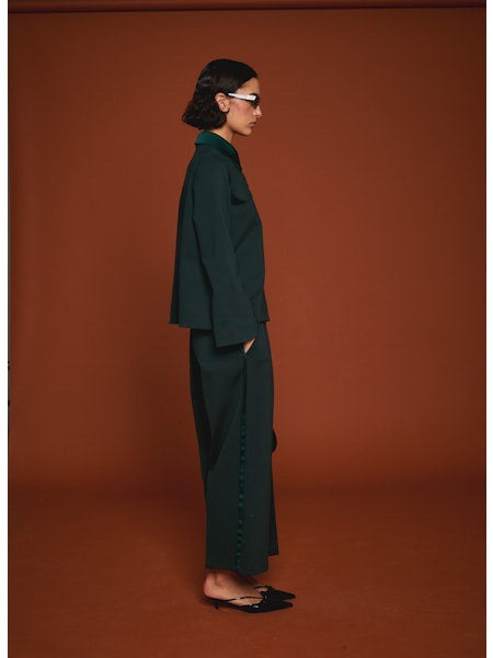 Facing away from the camera, a person wears MILVA’s MILVA MI - TUXEDO DRESS PANT in dark green with cropped legs and black heels, holding a black handbag. The background is solid brown.