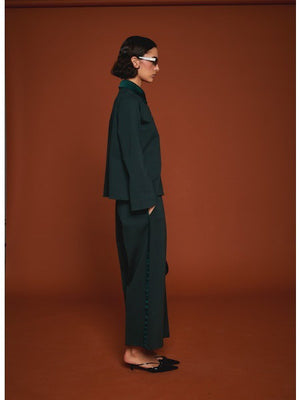 Facing away from the camera, a person wears MILVA’s MILVA MI - TUXEDO DRESS PANT in dark green with cropped legs and black heels, holding a black handbag. The background is solid brown.