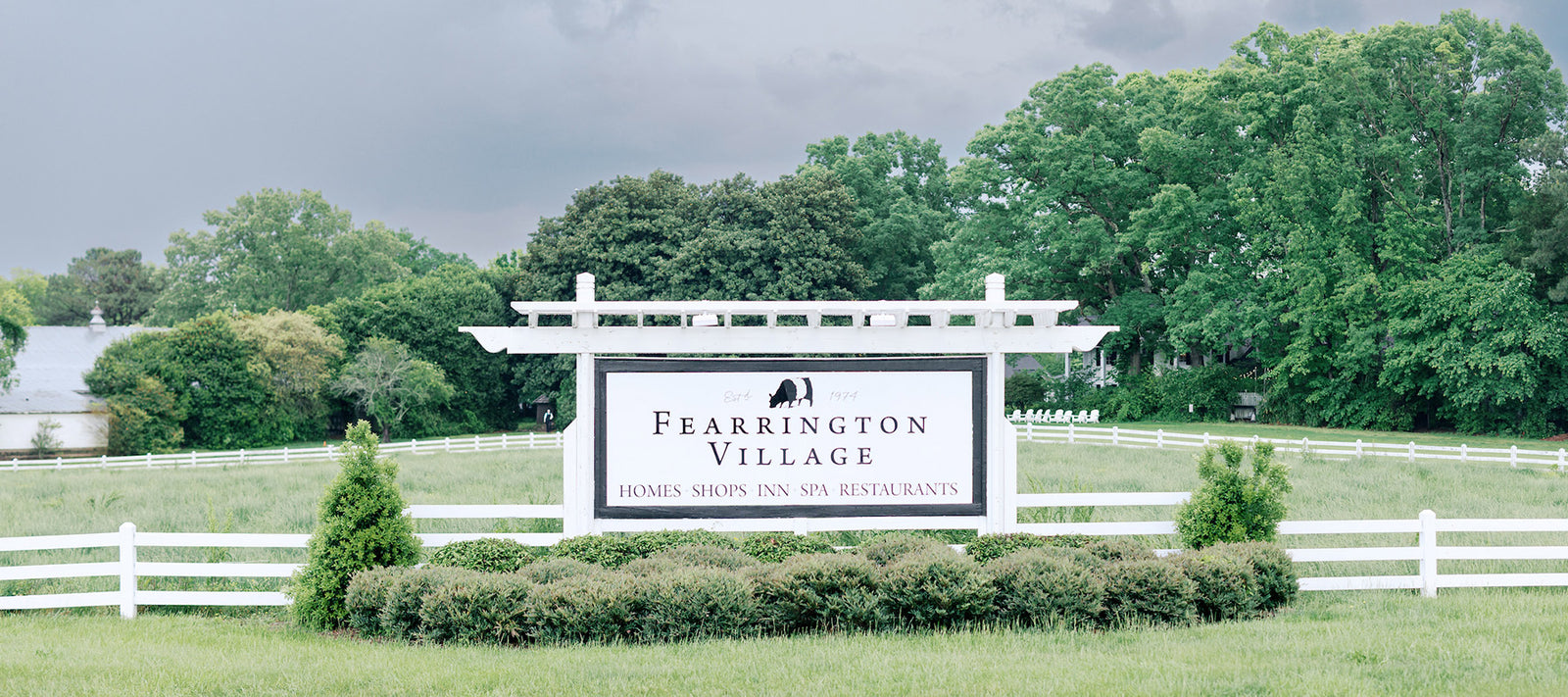 A white sign for Fearrington Village stands in front of a white fence, surrounded by green grass and trees under a cloudy sky. The sign advertises homes, shops, an inn, a spa, and restaurants.