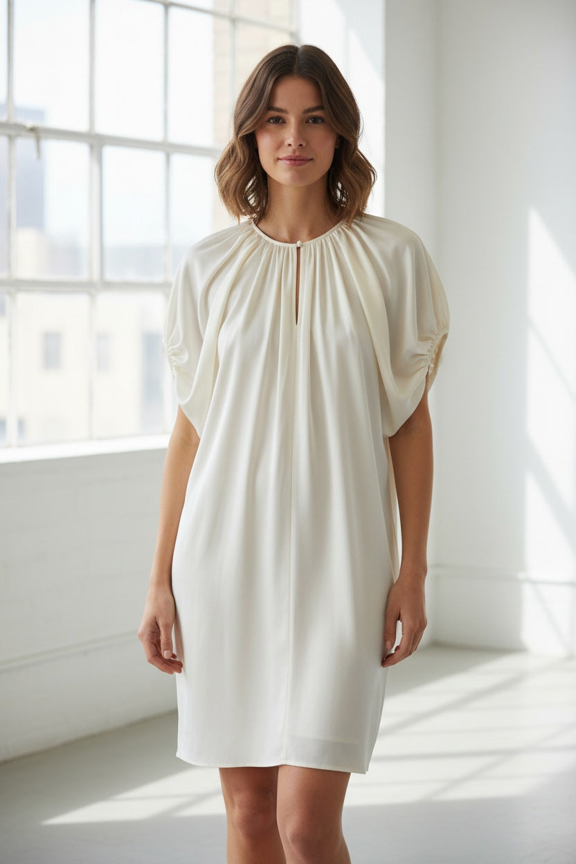 A woman stands indoors by a large window, dressed in the MOMONI - OLIVIA DRESS from MOMONI. The elegant white dress features gathered sleeves reminiscent of silk satin crepe. She pairs it with brown ankle boots, while the minimal background includes a gray wall and a partially visible plant.