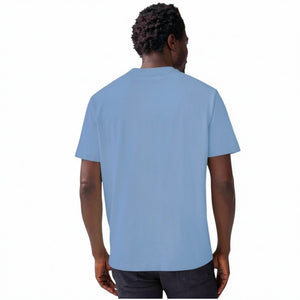 A man with short curly hair wears the MACK WELDON Heavyweight Slub Cotton Pocket T-Shirt in Dean Blue with dark pants, standing against a plain white background.