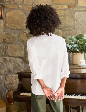 A person with curly hair, dressed in the FRANK & EILEEN OONA Continuous Sleeve Button Up Shirt in Superluxe Cotton White, and relaxed-fit green pants, stands facing away from the camera in front of a piano with their hands clasped behind their back. A stone wall and plants provide the backdrop.