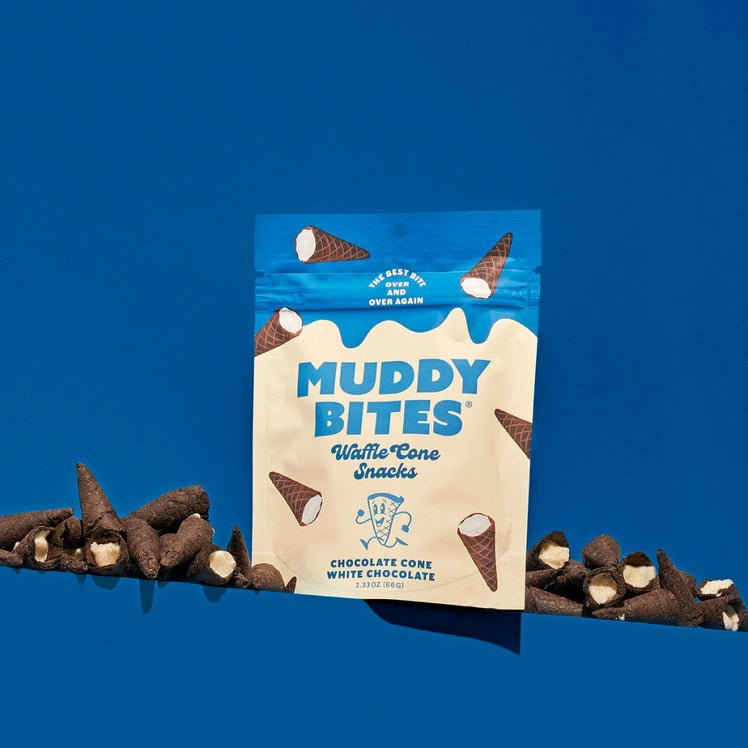 A person dressed in a light blue outfit sits on a plush, royal blue rug holding a bag of Muddy Bites Cookies &amp; Cream Waffle Cones in one hand and a small white chocolate waffle cone snack in the other. The beige and blue bag features an illustration of chocolate-dipped waffle cones.