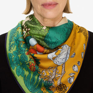 A woman wears the INOUI EDITIONS - QUATRE SAISON MODAL SILK SCARF by INOUI, featuring vibrant green, yellow, and orange patterns of trees, flowers, and abstract designs. Only her lower face, neck, and upper chest are visible. Made in India.