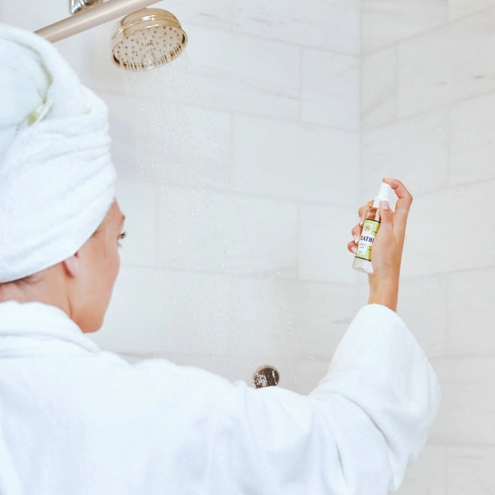 Six small bottles labeled Breathe, Boost, Unwind, Sleep, Detox, and Recover—each filled with essential oil shower mists like uplifting lemon and ginger—are arranged on white shelves in a tiled shower niche with a geometric gray and white pattern. Create your own aromatherapy steam room right at home.