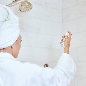 Six small bottles labeled Breathe, Boost, Unwind, Sleep, Detox, and Recover—each filled with essential oil shower mists like uplifting lemon and ginger—are arranged on white shelves in a tiled shower niche with a geometric gray and white pattern. Create your own aromatherapy steam room right at home.