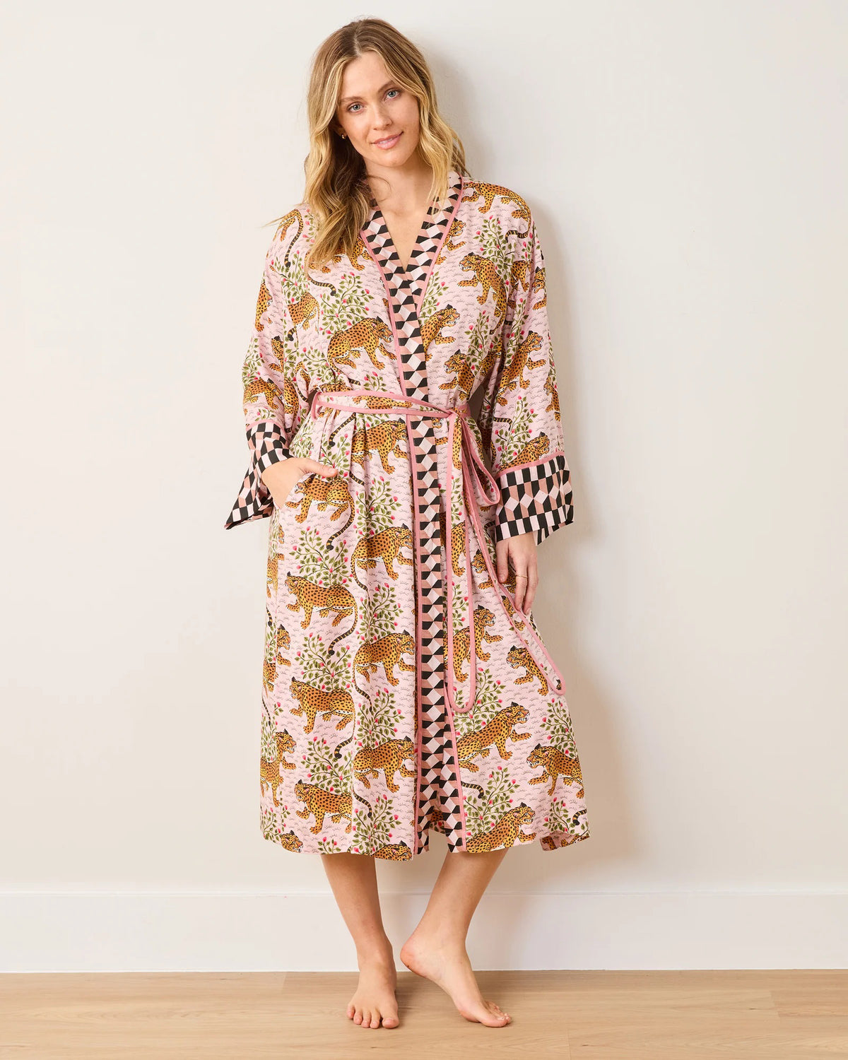 A person stands barefoot against a plain background, wearing the PRINTFRESH - SATIN ROBE BAGHEERA by PRINTFRESH. The eco-friendly viscose robe showcases a colorful leopard and foliage design, inspired by leopard print pajamas, with checkered trimmings and a matching belt. They have long, wavy hair and a relaxed expression.