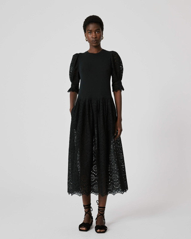 A person stands against a plain background wearing the SFIZIO - BRODERIE ANGLAISE MIDI DRESS by SFIZIO, which features elbow-length puffed sleeves and a midi-length skirt adorned with intricate lace patterns. They are also wearing black ankle-strap sandals.