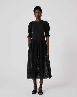 A person stands against a plain background wearing the SFIZIO - BRODERIE ANGLAISE MIDI DRESS by SFIZIO, which features elbow-length puffed sleeves and a midi-length skirt adorned with intricate lace patterns. They are also wearing black ankle-strap sandals.