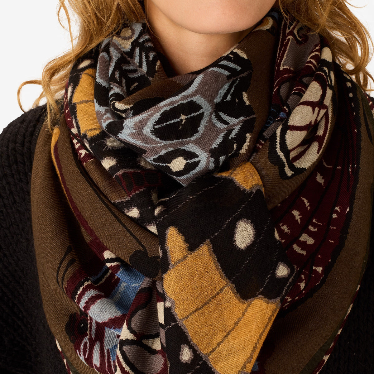 The INOUI EDITIONS - PAPILLONS WOOL SCARF IN KHAKI by INOUI is a square scarf made in India, featuring an intricate and colorful butterfly pattern in brown, blue, yellow, and white hues on a tan background. Measuring 130 x 130 cm, it provides generous coverage and stylish versatility for any occasion.
