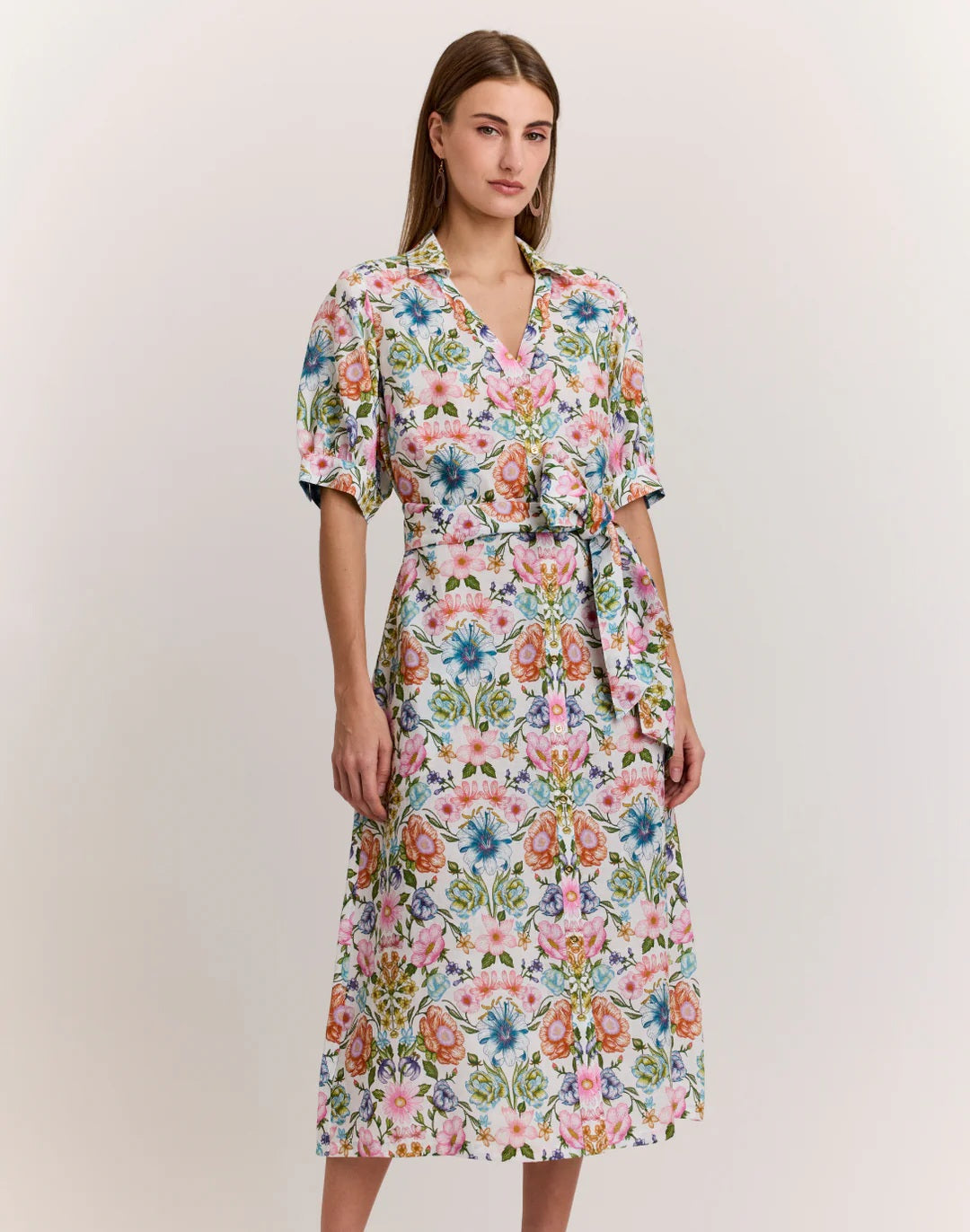 A woman with straight brown hair stands sideways, wearing the HINSON WU Luca Elbow Sleeve Linen Blend Dress in Botanique, a light floral midi dress with short sleeves and a waist tie, set against a plain light background.