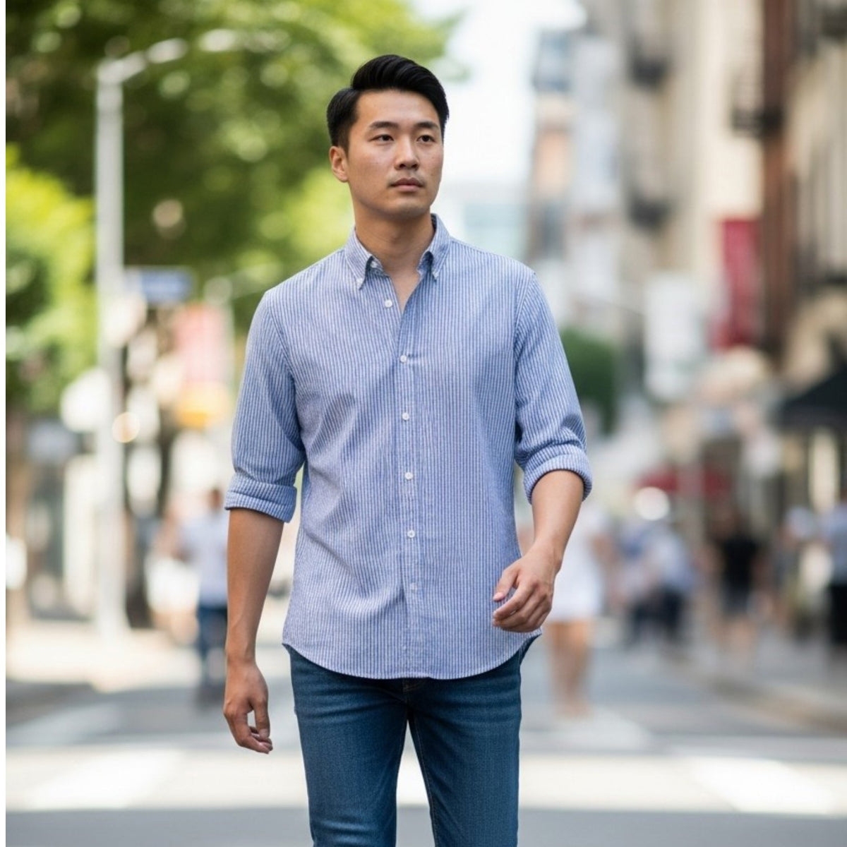 A man in a FRANK &amp; EILEEN Finbar Men's Button Up Shirt in Blue Stripes, blue jeans, and white sneakers walks confidently down a sunny, tree-lined cobblestone street.
