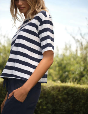 A person stands outdoors amidst green bushes and trees on a sunny day, wearing the FRANK & EILEEN Perry NYC Crewneck Tee in Vintage Jersey Nautical Stripe—a navy and white striped short-sleeve shirt crafted from 100% cotton—paired with dark pants.