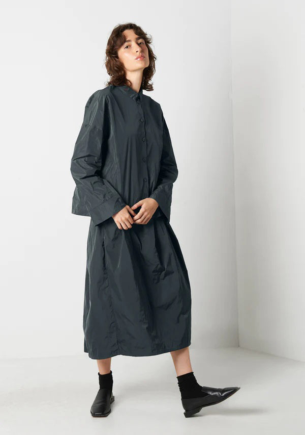 A person with shoulder-length wavy hair stands against a plain white background. They are wearing a dark gray button-up shirt, a matching KATHARINA HOVMAN - EASY SKIRT by KATHARINA HOVMAN, black socks, and black shoes. Their hands are casually placed in their pockets.