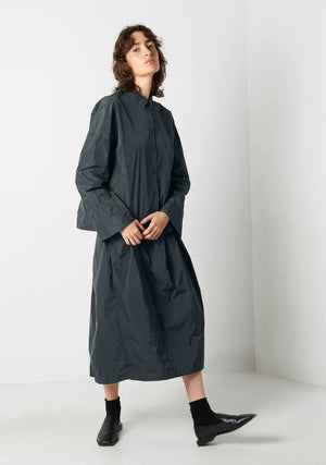 A person with shoulder-length wavy hair stands against a plain white background. They are wearing a dark gray button-up shirt, a matching KATHARINA HOVMAN - EASY SKIRT by KATHARINA HOVMAN, black socks, and black shoes. Their hands are casually placed in their pockets.