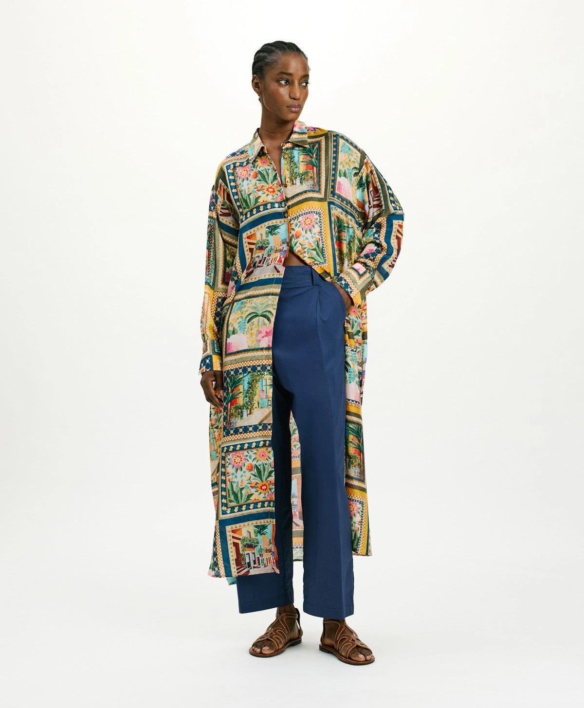 A person wears the MOMONI JAU DRESS, a colorful, artistic printed shirt dress, over blue high-waisted trousers against a plain backdrop. Brown sandals complete the effortless style as they gaze sideways.