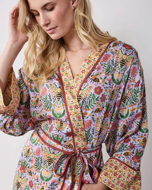 A woman wears a PRINTFRESH - BEE TAPESTRY ECO SATIN ROBE IN LAVENDER, featuring a lavender floral-print with a repeating pattern of bees, flowers, and vines with a contrasting yellow and red patterned trim.