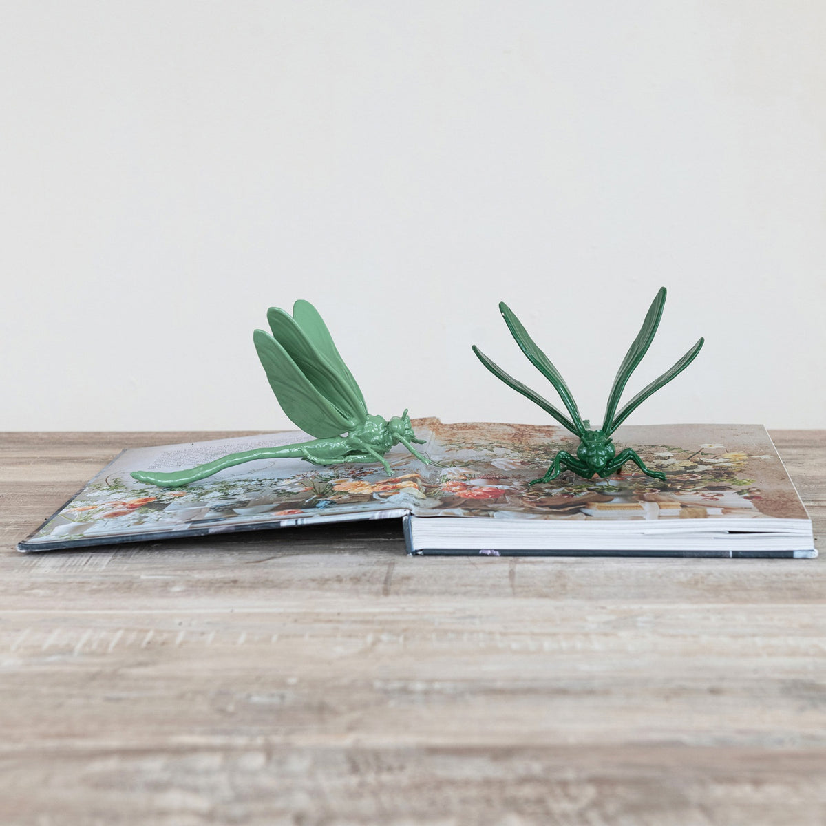 A pop-up book on a wooden surface showcases two large green CAST IRON DRAGONFLY sculptures by CREATIVE COOP, emerging from its pages against a plain light background.