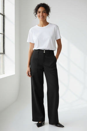 A person wears HINSON WU's KELLY TRAVELER'S TWILL PANT in dark green with front pockets and gold buttons, styled with a white top and black pointed-toe shoes. Photo is cropped from shoulders down, highlighting this travel-ready look.