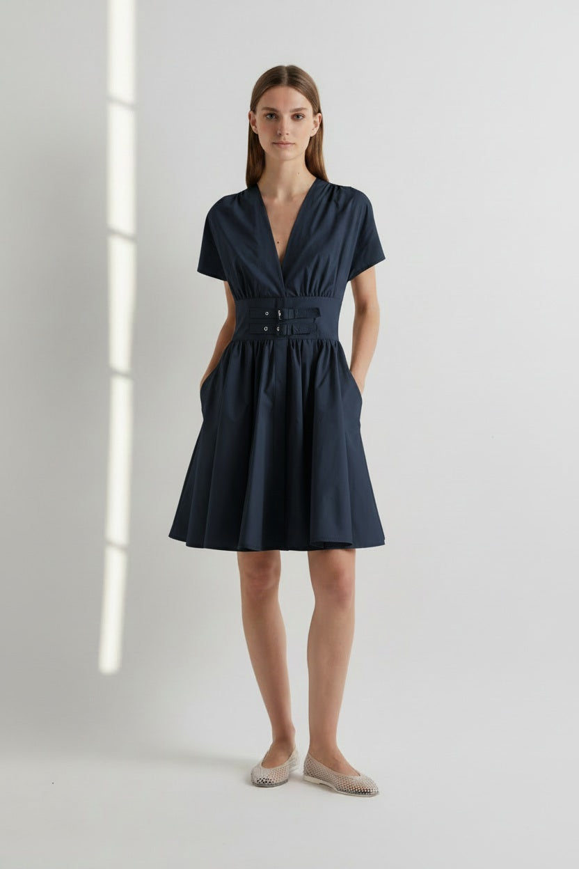 A woman stands against a white wall with light streaming in from the left, wearing the MEIMEIJ Short Cottone Dress with Front Buckle in Dark Navy by MEIMEIJ—a short-sleeved, cotton dress featuring a belted waist, pleated skirt, subtle corset-inspired details, and handy pockets. She completes the look with beige flats.