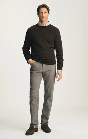 A man stands against a plain background wearing a dark brown sweater layered over a light collared shirt, paired with 34 HERITAGE - COURAGE STRAIGHT LEG PANTS IN STONE DIAGONAL by 34 HERITAGE, and brown shoes. One hand rests in his pocket and he maintains a neutral expression.