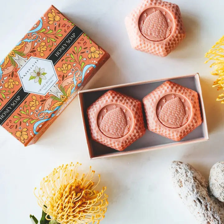 Experience the BAUDELAIRE Orange Blossom Honey Soap 3.5 oz, 2 Bar Gift Box—a beautifully presented set featuring two hexagonal orange soaps nestled in an open decorative box adorned with floral designs and a central bee illustration. This triple-milled soap by BAUDELAIRE offers a luxurious lather and a delicate orange blossom honey fragrance, making it an ideal indulgence for yourself or a thoughtful gift for someone special.