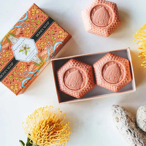 Experience the BAUDELAIRE Orange Blossom Honey Soap 3.5 oz, 2 Bar Gift Box—a beautifully presented set featuring two hexagonal orange soaps nestled in an open decorative box adorned with floral designs and a central bee illustration. This triple-milled soap by BAUDELAIRE offers a luxurious lather and a delicate orange blossom honey fragrance, making it an ideal indulgence for yourself or a thoughtful gift for someone special.