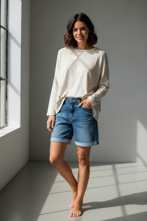 A woman stands barefoot in sunlight, smiling with one hand in her pocket while wearing the MERSEA - CATALINA RUSTIC TEE IN BONE paired with blue denim shorts, in a minimalist room.