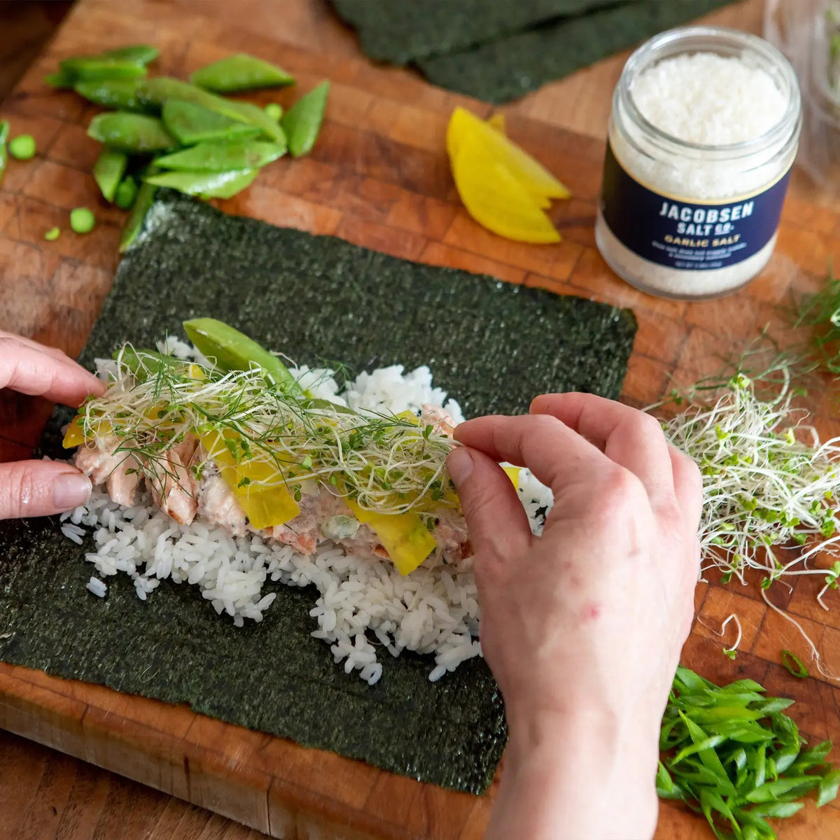 A glass jar of Jacobsen Salt Co. Garlic Salt Infused Sea Salt, featuring a metal lid and a black-and-gold label, filled with coarse garlic-infused sea salt made from premium kosher salt.