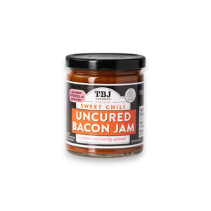 A jar of TBJ GOURMET Sweet Chili Uncured Bacon Jam sports a sleek black and orange label with the phrase "a warm and smoky spread" on the front. Sealed with a metal lid, this gluten-free jam is ideal for culinary adventures.