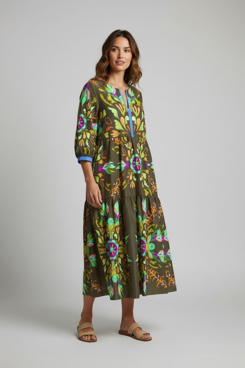 A woman wears the FRANCO FERRARI - SILK TIERED MIDI DRESS, a long sleeve olive green silk piece with colorful floral and abstract prints. She smiles softly, has wavy hair, beige sandals, and stands against a plain light gray background.