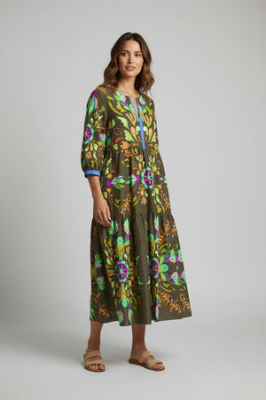 A woman wears the FRANCO FERRARI - SILK TIERED MIDI DRESS, a long sleeve olive green silk piece with colorful floral and abstract prints. She smiles softly, has wavy hair, beige sandals, and stands against a plain light gray background.