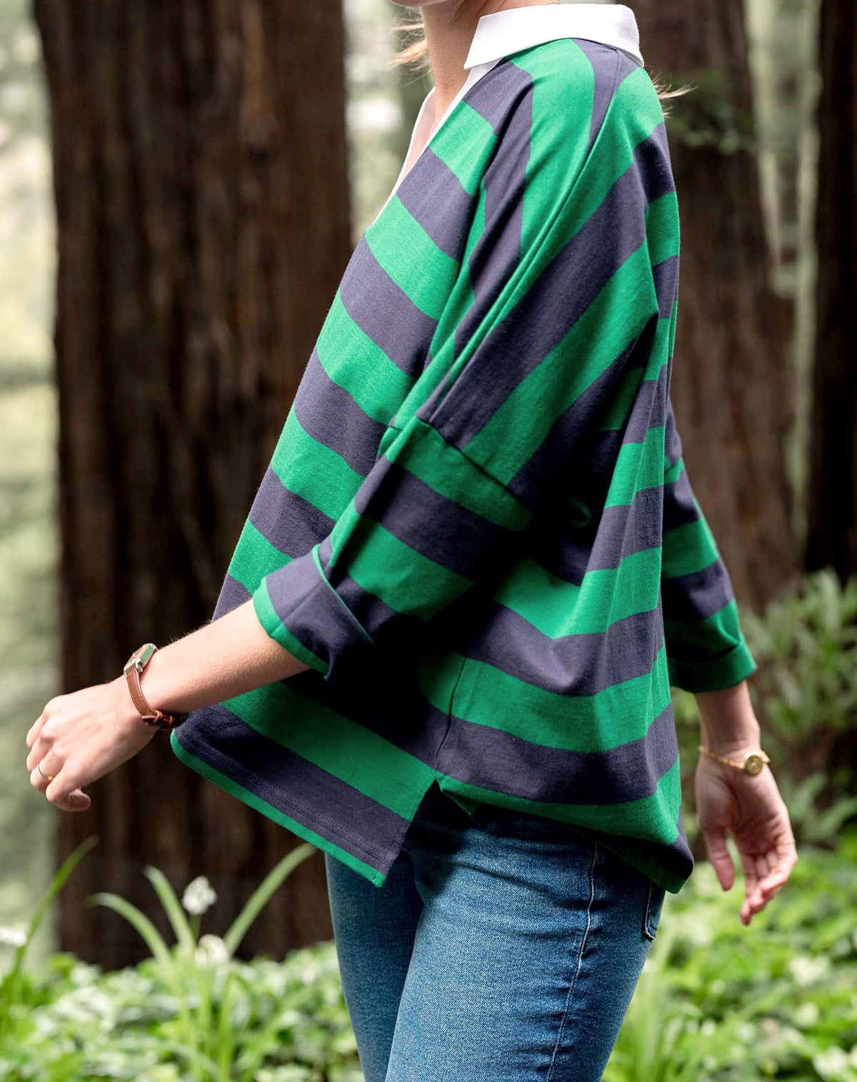 A person wears the MERSEA Amelia Rugby Tee in navy green stripe, featuring a white collar, and blue jeans. They stand outdoors with hands in pockets, green foliage visible behind them.